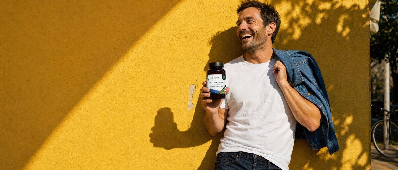 Lachender Mann an einer gelben Hauswand mit einem Glas Echt Vital Magnesium Komplex in der Hand